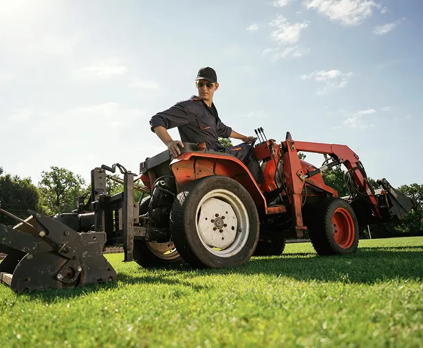 A Kickin’ Grass crew member driving an orange compact tractor with a front loader across a mown lawn